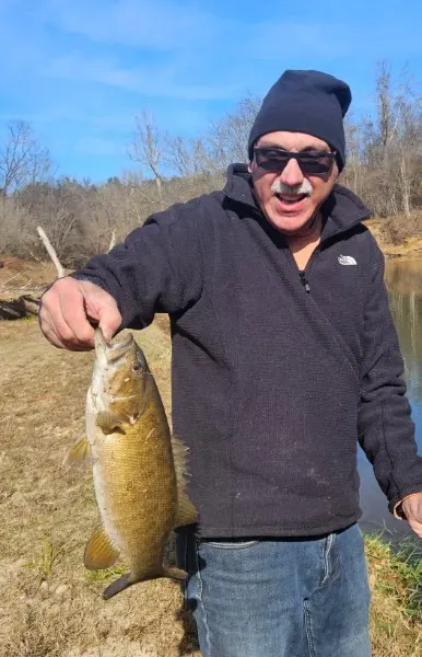 A smiling camper in sunglasses and a black knit cap holding a large bass caught from Yadkin River.