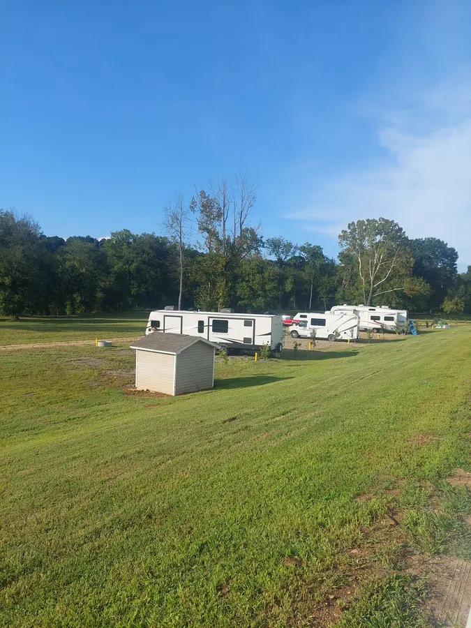 Several RVs and vehicles parked at campsites across a sunny, grassy campground, viewed from a nearby hillside.