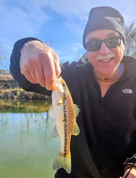 A smiling camper in sunglasses and a black knit cap holding a small bass caught from Yadkin River.