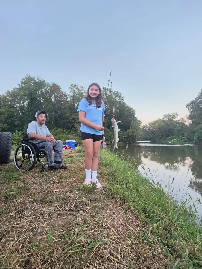 A young angler holding a fish beside Yadkin River while two companions relax nearby, sharing great fishing and fun at Hillbilly Campground.